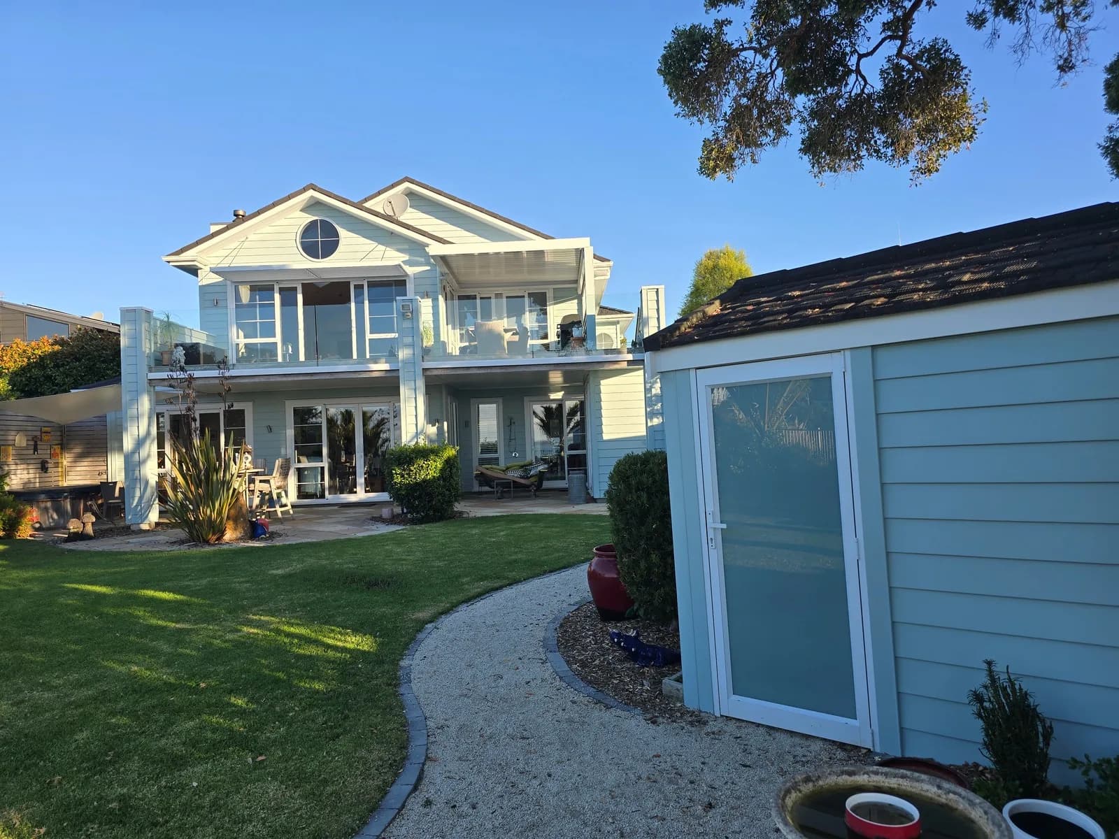 Two-storey pale blue house with white balconies and a matching blue garden shed beside a curved gravel path