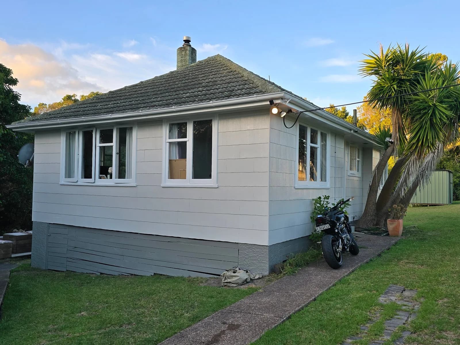 Single-storey bungalow with white weatherboards, white windows and a dark grey base, with cabbage trees beside it