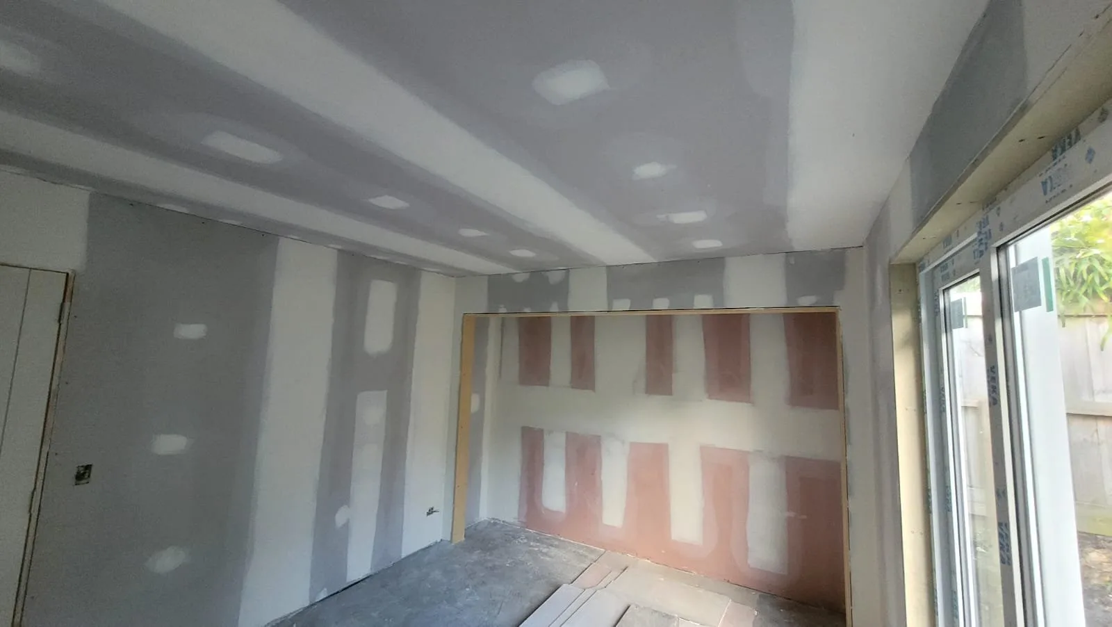 New plasterboard lining on the walls and ceiling of a room, with pink fire-rated board on the back wall and the first coat of stopping over the joints and screws