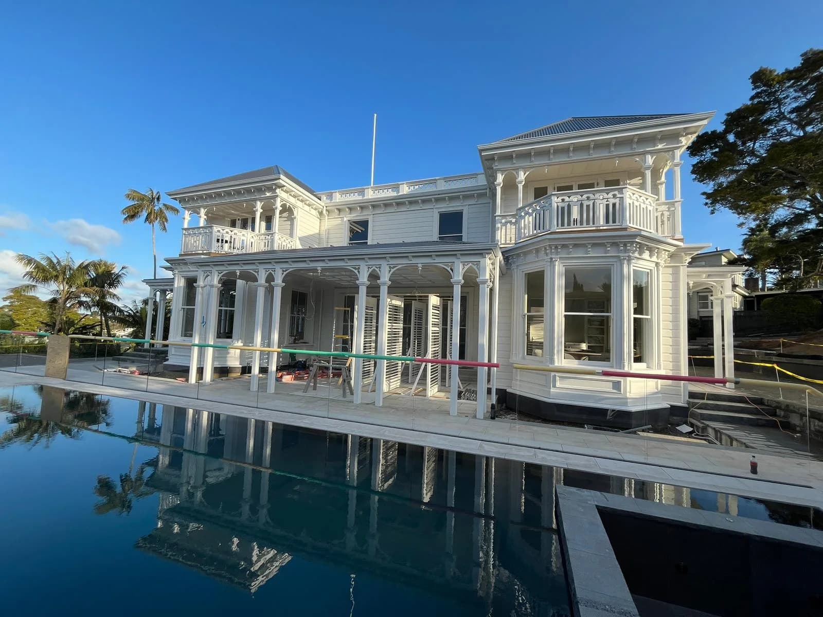 Two-storey white weatherboard villa with ornate verandas and balconies, fully repainted, reflected in the swimming pool in front of it